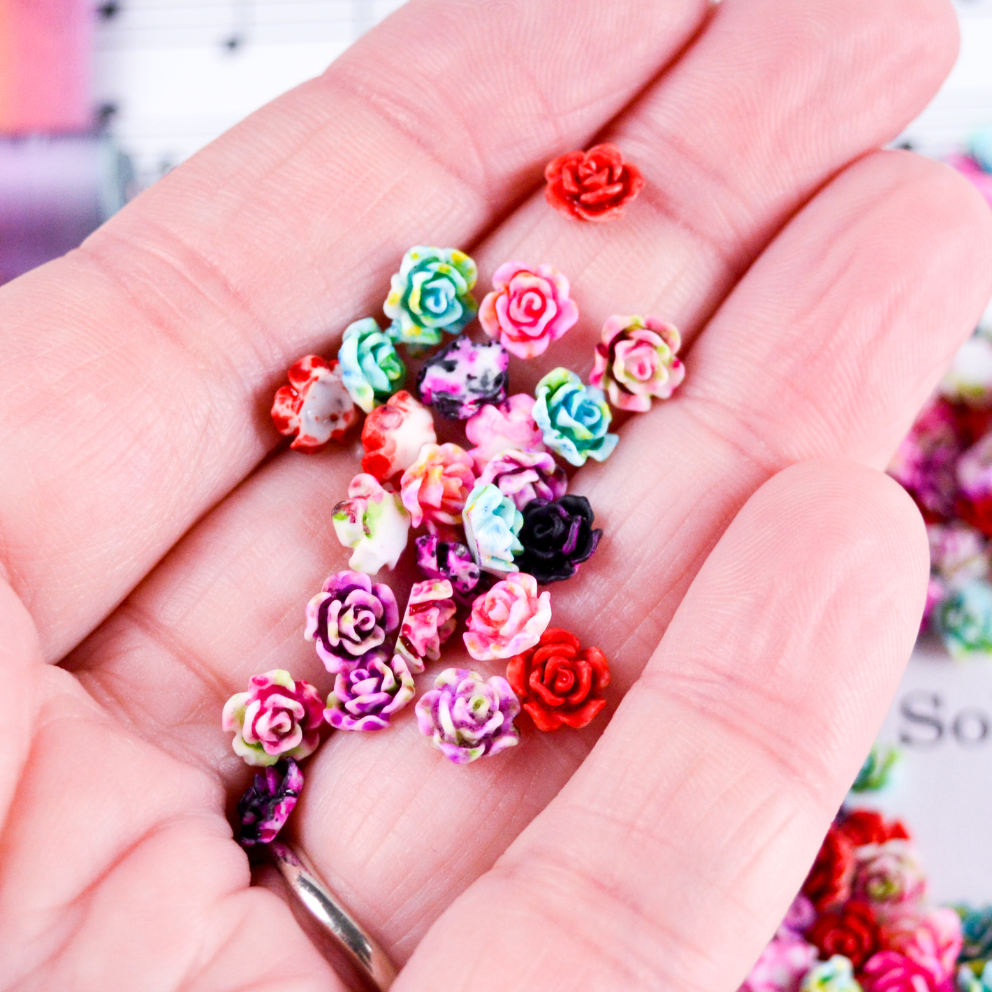 Closeup image of an open hand, holding dozens of tiny 6mm rose cabochons in splatter painted pinks, greens, red, black and purples.